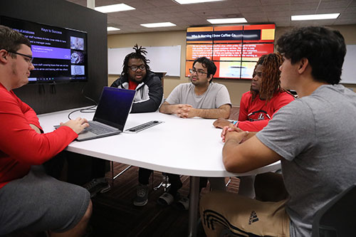 A group of diverse students sit in around a table discussing business strategies.