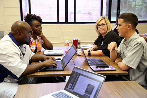 A group of students converse at a table filled with laptops and notebooks