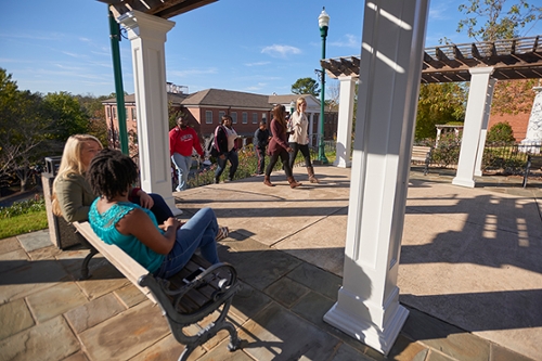 Two students on a bench in the foreground chat as other students crest the stairs to the patio outside the college library.