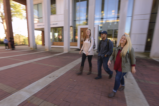 Three smiling students walk past the library on the plaza.